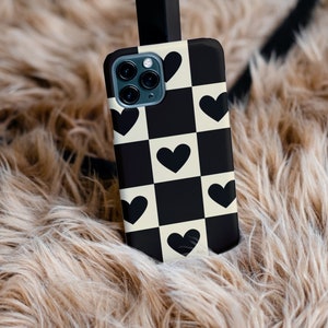 May include: A black and white chequered phone case with black hearts. The case is on a light brown fluffy surface.