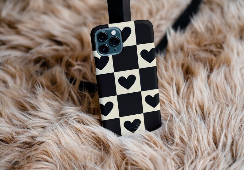May include: A black and white chequered phone case with black hearts. The case is on a light brown fluffy surface.