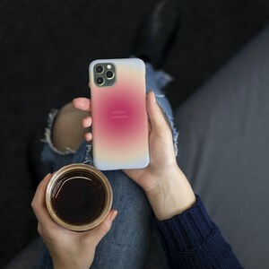 Pu&ograve; includere: Una persona tiene in mano una custodia per iPhone bianca con un gradiente di rosa, arancione e giallo. La custodia ha il testo "positivit&agrave;" e "il tuo potere" stampato su di essa. La persona tiene anche una tazza di caff&egrave;.