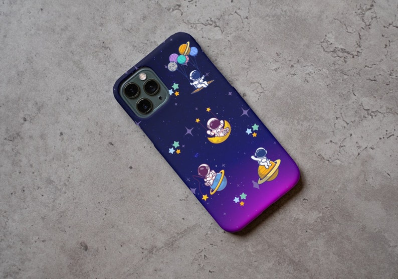 Peut inclure: Une coque de t&eacute;l&eacute;phone de couleur violette et bleue sur le th&egrave;me de l'espace. Elle pr&eacute;sente un fond de galaxie avec des &eacute;toiles et des plan&egrave;tes, et quatre astronautes flottant dans l'espace.
