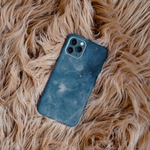 May include: A blue phone case with a galaxy print and a white ring design. The case is lying on a brown fuzzy surface.