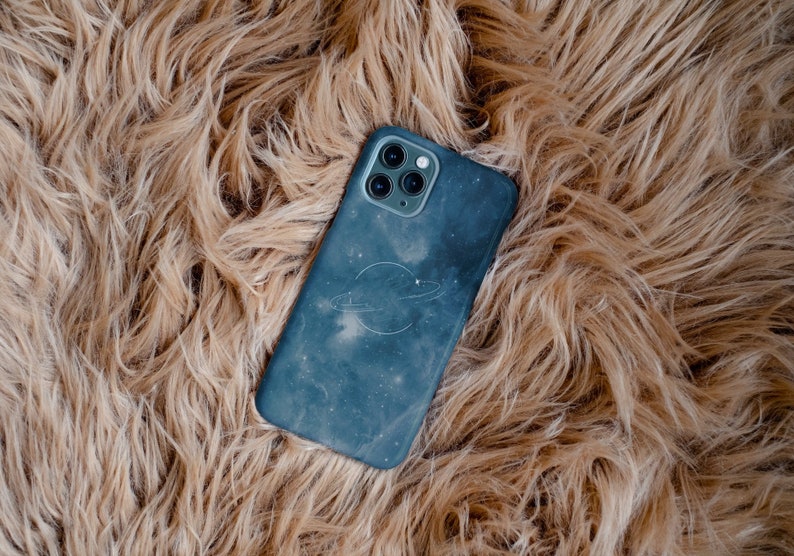 May include: A blue phone case with a galaxy print and a white ring design. The case is lying on a brown fuzzy surface.