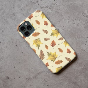 May include: A white phone case with a pattern of brown and yellow leaves. The case has a camera lens cutout.