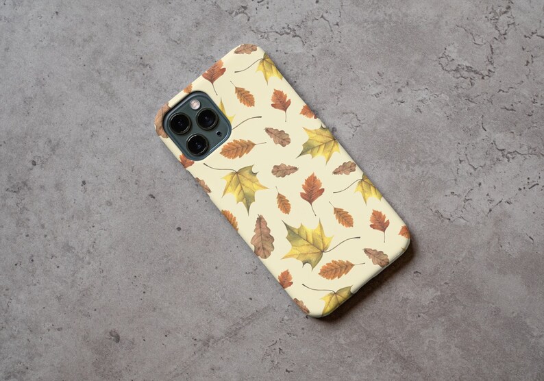 May include: A white phone case with a pattern of brown and yellow leaves. The case has a camera lens cutout.