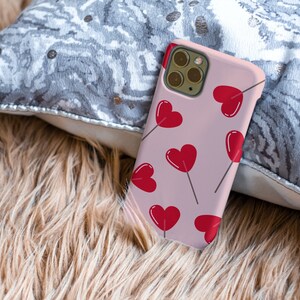 May include: A pink phone case with a pattern of red hearts on sticks. The case has a camera lens cutout and a white border.