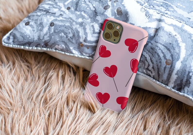 May include: A pink phone case with a pattern of red hearts on sticks. The case has a camera lens cutout and a white border.