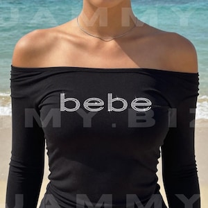 May include: A black long-sleeved top with an off-the-shoulder neckline featuring the word "bebe" in a sparkling rhinestone font across the chest. The garment is plain black fabric with a fitted silhouette and long sleeves.