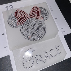 Pode incluir: Um fundo branco com um design de strass de uma cabeça de rato com um laço vermelho e a palavra "GRACE" em strass.