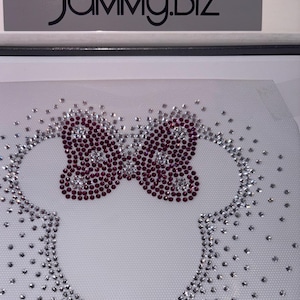 Può includere: Un tessuto a rete bianco con un design di testa di topo dei cartoni animati con un fiocco rosa fatto di strass. Il design è per l'applicazione con il ferro da stiro. Il testo "JAMMY.BIZ" è nella parte superiore dell'immagine.