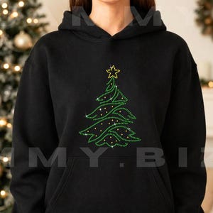 May include: A black hooded sweatshirt featuring a central graphic of a Christmas tree made of green and yellow rhinestones. The tree design includes a yellow star at the top and scattered yellow accents throughout the green branches.