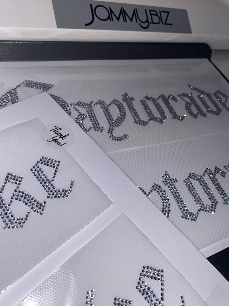 Puede incluir: M&uacute;ltiples transferencias de diamantes de imitaci&oacute;n termoadhesivos sobre hojas de soporte blancas. Los dise&ntilde;os presentan cristales de color plateado dispuestos en letras de estilo g&oacute;tico. Una hoja muestra el texto "Gatorade" y otra una pegatina "Thank You!".