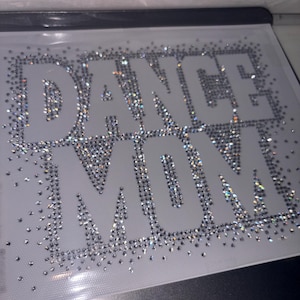 May include: A close-up of a white fabric with the words "Dance Mom" spelled out in rhinestones. The rhinestones are clear and sparkly.