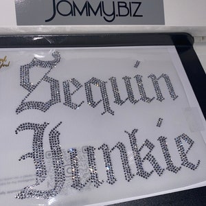 Puede incluir: Una hoja de transferencia de pedrer&iacute;a con el texto Sequin Junkie en una fuente g&oacute;tica plateada. Las letras est&aacute;n compuestas por peque&ntilde;os cristales transparentes reflectantes dispuestos sobre un soporte adhesivo transparente. La marca JAMMY.BIZ es visible en la parte superior.