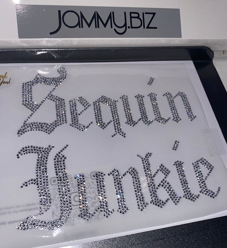 Puede incluir: Una hoja de transferencia de pedrer&iacute;a con el texto Sequin Junkie en una fuente g&oacute;tica plateada. Las letras est&aacute;n compuestas por peque&ntilde;os cristales transparentes reflectantes dispuestos sobre un soporte adhesivo transparente. La marca JAMMY.BIZ es visible en la parte superior.