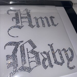Puede incluir: Una hoja de transferencia de diamantes de imitaci&oacute;n con el texto "Hmc Baby" en una fuente g&oacute;tica blackletter. Las letras est&aacute;n compuestas por peque&ntilde;os cristales claros y reflectantes sobre un soporte adhesivo blanco para aplicaci&oacute;n t&eacute;rmica.