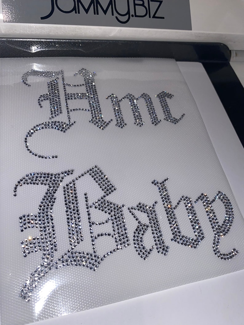 Puede incluir: Una hoja de transferencia de diamantes de imitaci&oacute;n con el texto "Hmc Baby" en una fuente g&oacute;tica blackletter. Las letras est&aacute;n compuestas por peque&ntilde;os cristales claros y reflectantes sobre un soporte adhesivo blanco para aplicaci&oacute;n t&eacute;rmica.