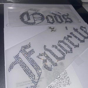 Puede incluir: Dos transferencias de diamantes de imitaci&oacute;n termoadhesivos sobre un soporte adhesivo transparente. La transferencia superior muestra la palabra "God's" en fuente g&oacute;tica y la inferior la palabra "Favorite" en el mismo estilo. Cristales claros y brillantes.