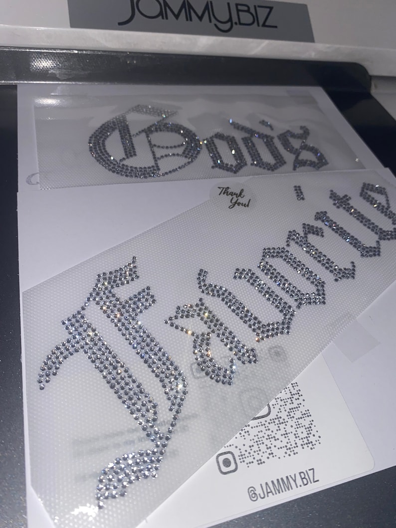 Puede incluir: Dos transferencias de diamantes de imitaci&oacute;n termoadhesivos sobre un soporte adhesivo transparente. La transferencia superior muestra la palabra "God's" en fuente g&oacute;tica y la inferior la palabra "Favorite" en el mismo estilo. Cristales claros y brillantes.