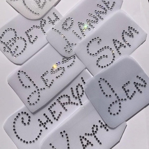 Puede incluir: Un conjunto de nueve parches de tela blanca con varios nombres escritos en pedrería. Los nombres incluyen "Leah", "Breanna", "Grace", "Chloe", "Carolyn", "Olivia" y "Karm"
