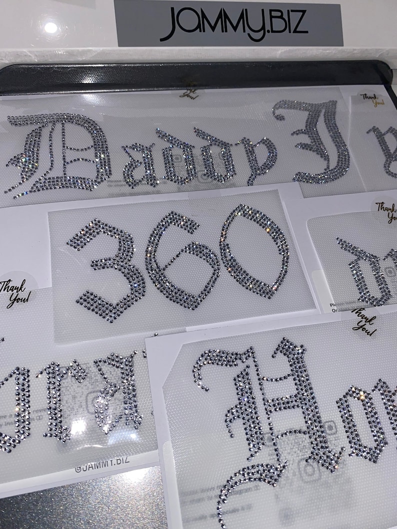 Puede incluir: M&uacute;ltiples transferencias de diamantes de imitaci&oacute;n para planchar con cristales de color plateado dispuestos en letras y n&uacute;meros de estilo g&oacute;tico. Los dise&ntilde;os incluyen el texto "Daddy", el n&uacute;mero "360" y otras palabras parciales en l&aacute;minas adhesivas transparentes con etiquetas "Thank You".