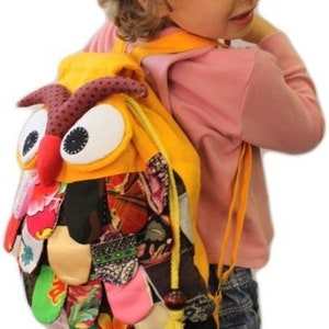 May include: A yellow owl-shaped backpack with large white eyes, a red beak, and brown details. The backpack has a patchwork design with various colourful fabric pieces. The straps are yellow, and the backpack is worn by a child.