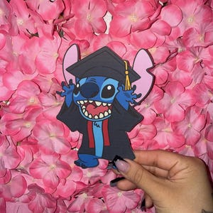 Op de afbeelding: Een blauw stripfiguur met een afstudeerhoed en toga, met de armen enthousiast omhoog. Het personage heeft grote oren en een brede grijns. De achtergrond is een dichte opstelling van roze bloemen.