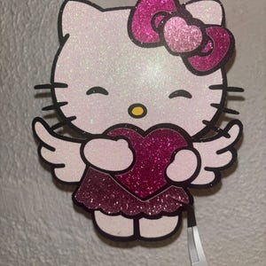 Pode incluir: Uma figura de Hello Kitty de glitter branco e rosa com asas de anjo segurando um coração de glitter rosa.