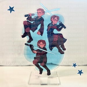 May include: A clear acrylic stand featuring three illustrations of a character with a lightsaber. The character is depicted in a brown and maroon outfit with a lightsaber. The background is light blue with star stickers.