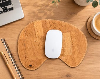 以下が含まれることがあります： 薄茶色のテクスチャ加工されたコルク製マウスパッド。曲線的で非対称な形状で、左下隅に小さな木のロゴが印刷されています。滑らかな白い楕円形のコンピュータマウスが、斑点のある表面を持つマウスパッドの中央に置かれています。