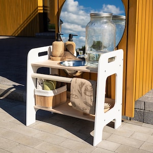 May include: A white wooden play kitchen with a wooden countertop, a stainless steel sink, and a large glass jar with water. The kitchen has a shelf with a basket, a towel, and a brush. A round mirror is in the background.