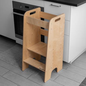 Op de afbeelding: Een lichtgekleurde houten keukentrap met twee treden en een bovenste plank. De kruk heeft uitgesneden handgrepen en is ontworpen om kinderen te helpen werkbladen te bereiken. De merknaam "GrowKids" is zichtbaar aan de voorkant.