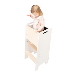 May include: A white wooden learning tower with a child standing on the top platform. The tower has a step ladder and a safety rail. The child is wearing a pink shirt and is holding a pink toy.