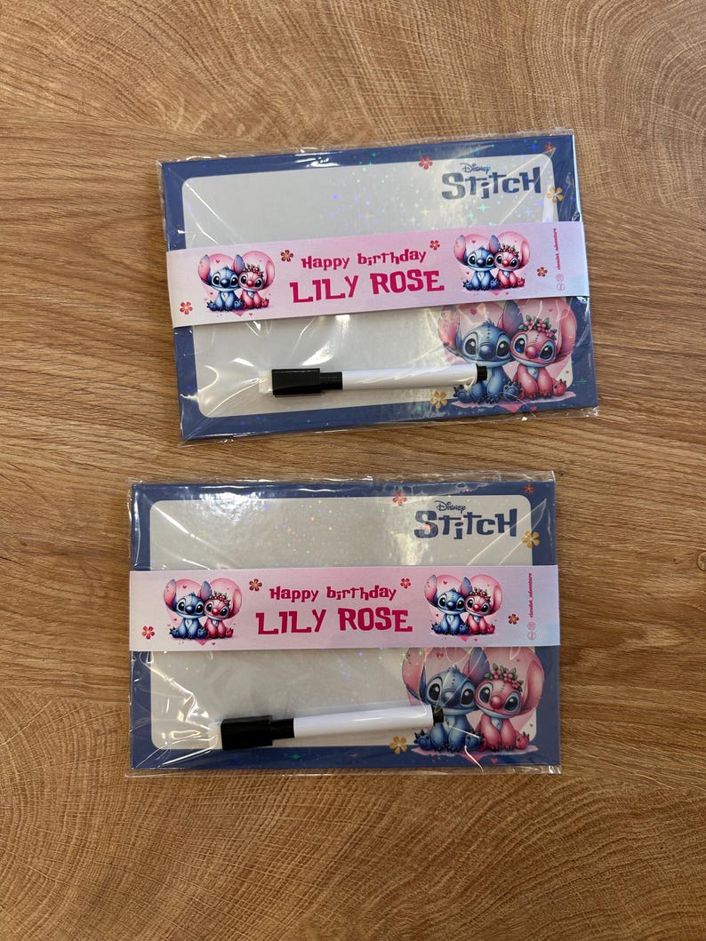 Puede incluir: Dos pizarras blancas rectangulares de Disney Stitch envueltas en pl&aacute;stico transparente. Cada pizarra tiene una banda de papel rosa con el texto Happy birthday LILY ROSE e ilustraciones de Stitch y Angel. Incluye un marcador blanco peque&ntilde;o.