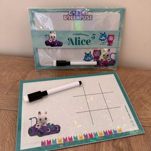 Puede incluir: Un set de pizarra blanca borrable con tem&aacute;tica de Gabby's Dollhouse que incluye un tablero con una cuadr&iacute;cula de tres en raya, un personaje en un coche morado y una fila de iconos de gatos de colores. Incluye un rotulador blanco borrable.