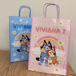Puede incluir: Dos bolsas de regalo rectangulares de papel con asas. Una bolsa es azul claro y la otra rosa claro. Ambas presentan una ilustraci&oacute;n de perro de dibujos animados, un arco&iacute;ris, margaritas blancas y el nombre VIVIANA con el n&uacute;mero 2 en la bolsa rosa.
