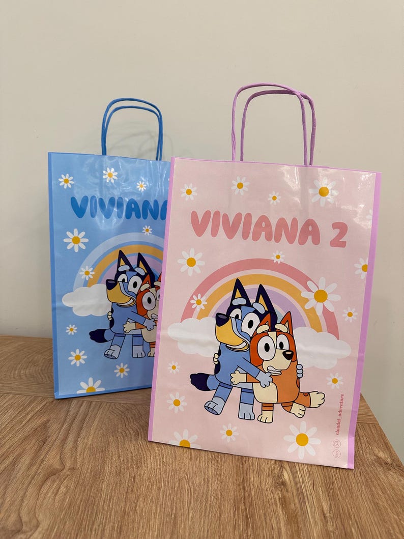 Puede incluir: Dos bolsas de regalo rectangulares de papel con asas. Una bolsa es azul claro y la otra rosa claro. Ambas presentan una ilustraci&oacute;n de perro de dibujos animados, un arco&iacute;ris, margaritas blancas y el nombre VIVIANA con el n&uacute;mero 2 en la bolsa rosa.