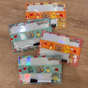 Puede incluir: Cuatro pizarras blancas rectangulares con la marca Pok&eacute;mon e ilustraciones de personajes. Cada pizarra est&aacute; envuelta en una banda de papel con el texto 'COMPLEANNO DI LEO', Charmander, Pikachu, Squirtle o Bulbasaur, e incluye un marcador borrable.