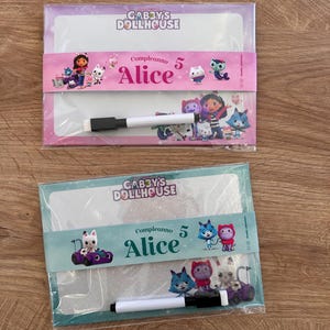 Puede incluir: Dos pizarras blancas rectangulares con personajes de Gabby's Dollhouse. La pizarra superior tiene una banda rosa con el texto "Compleanno 5 Alice" y un marcador. La inferior tiene una banda turquesa con el mismo texto y un marcador.