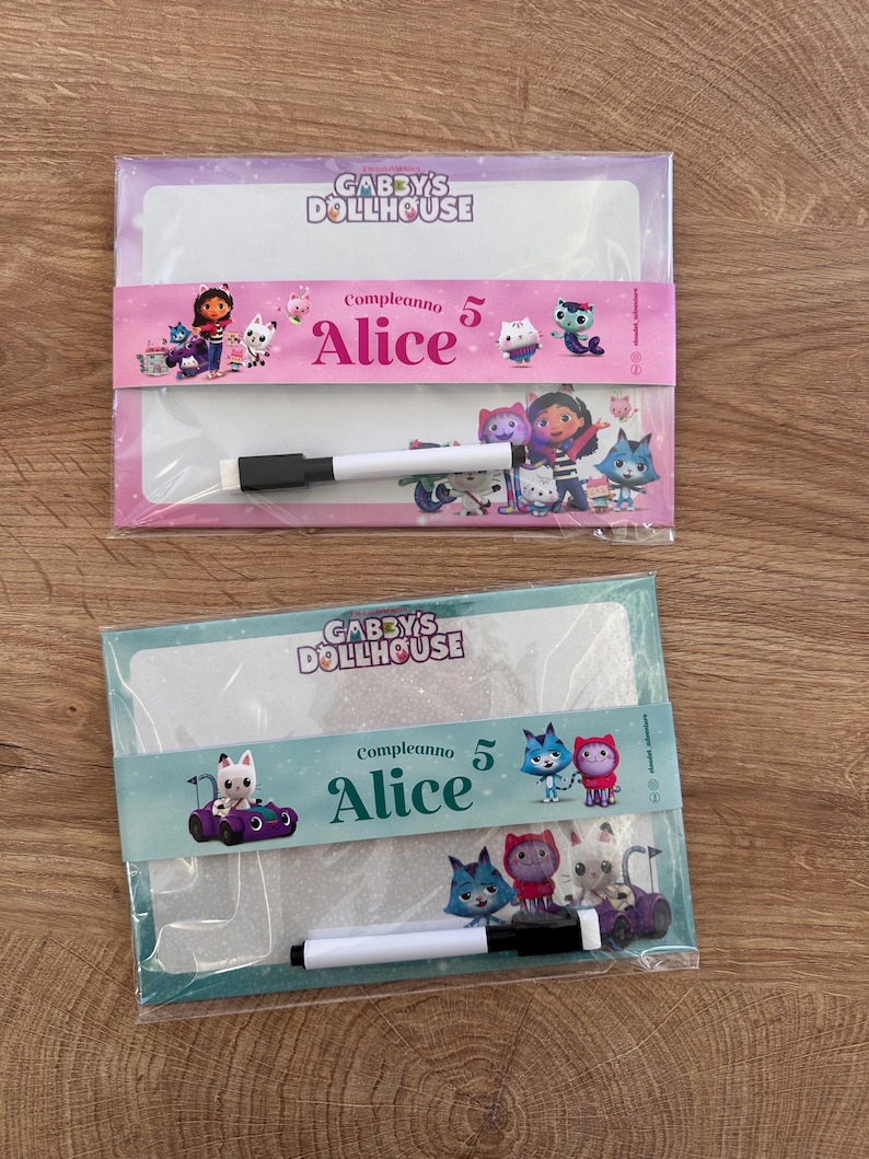 Puede incluir: Dos pizarras blancas rectangulares con personajes de Gabby's Dollhouse. La pizarra superior tiene una banda rosa con el texto "Compleanno 5 Alice" y un marcador. La inferior tiene una banda turquesa con el mismo texto y un marcador.