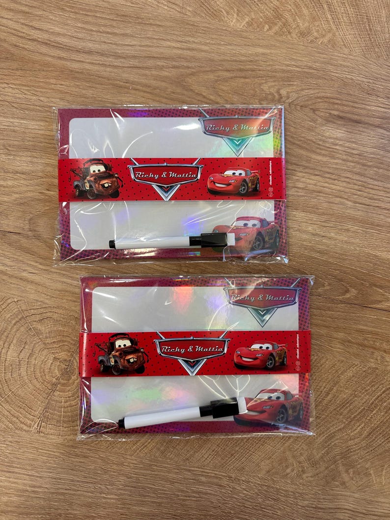 Puede incluir: Dos pizarras blancas rectangulares envueltas en pl&aacute;stico transparente, cada una con una banda roja con el texto Richy & Mattia e im&aacute;genes de coches de dibujos animados. Se incluye un peque&ntilde;o rotulador de pizarra blanco y negro con cada tabla.