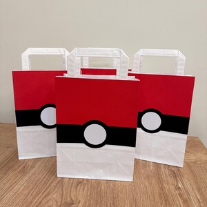 Puede incluir: Tres bolsas de regalo rectangulares de papel con un dise&ntilde;o inspirado en una Pok&eacute; Ball. Cada bolsa tiene una secci&oacute;n superior roja, una secci&oacute;n inferior blanca y una franja negra horizontal en el medio con un c&iacute;rculo blanco centrado.