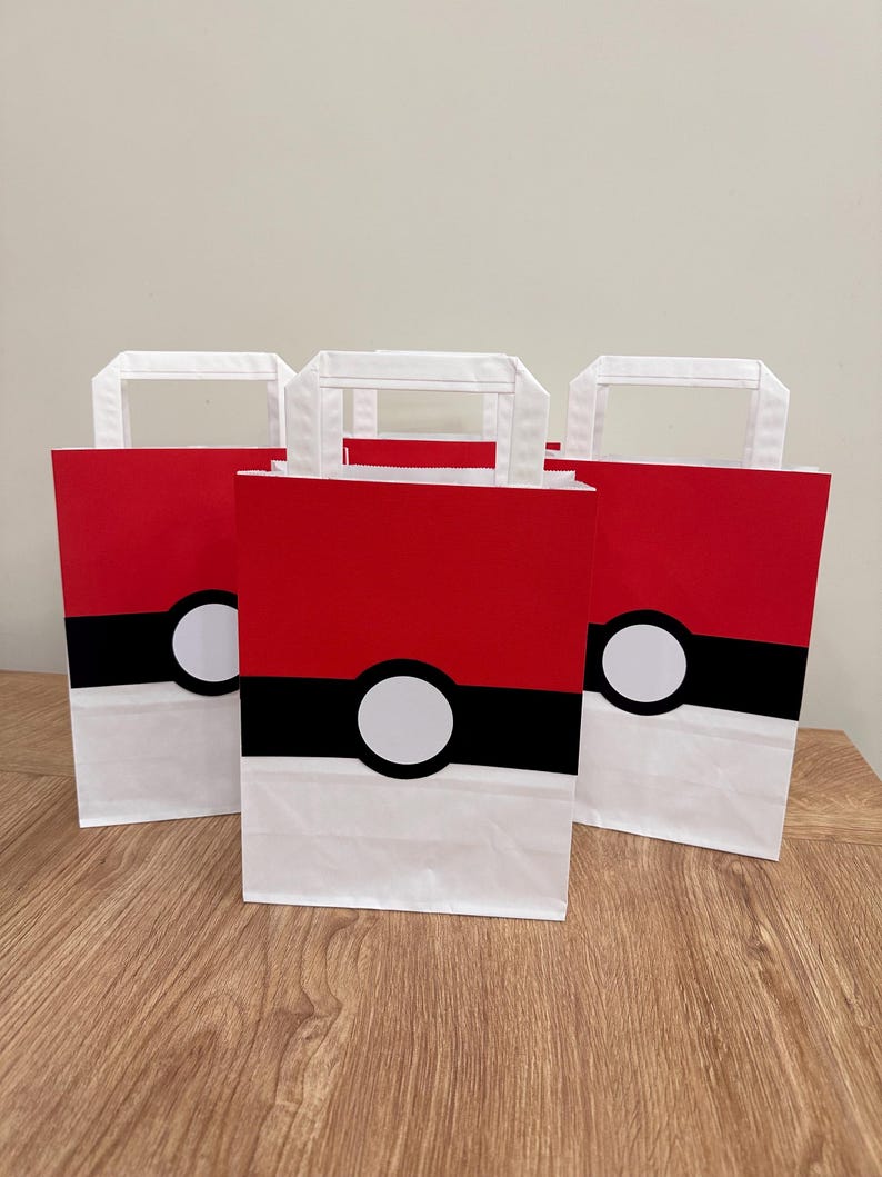 Puede incluir: Tres bolsas de regalo rectangulares de papel con un dise&ntilde;o inspirado en una Pok&eacute; Ball. Cada bolsa tiene una secci&oacute;n superior roja, una secci&oacute;n inferior blanca y una franja negra horizontal en el medio con un c&iacute;rculo blanco centrado.