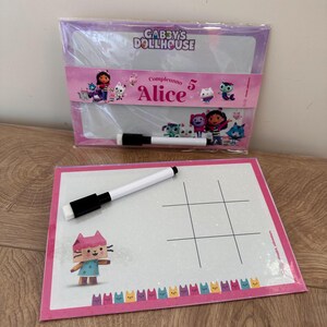Puede incluir: Dos pizarras blancas rectangulares de borrado en seco con gr&aacute;ficos de Gabby's Dollhouse. Una est&aacute; en un paquete de pl&aacute;stico con el texto "Gabby's Dollhouse" y "Compleanno 5 Alice". La otra tiene una cuadr&iacute;cula de tres en raya, un gato y dos rotuladores.