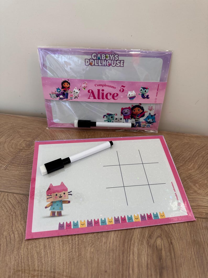 Puede incluir: Dos pizarras blancas rectangulares de borrado en seco con gr&aacute;ficos de Gabby's Dollhouse. Una est&aacute; en un paquete de pl&aacute;stico con el texto "Gabby's Dollhouse" y "Compleanno 5 Alice". La otra tiene una cuadr&iacute;cula de tres en raya, un gato y dos rotuladores.