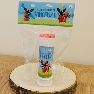 Può includere: Un flacone per bolle di sapone in un sacchetto di plastica trasparente con intestazione in cartoncino. L'etichetta riporta il testo "IL COMPLEANNO DI VINCENZO" e "2" con i personaggi Bing e Sula su sfondo verde e cielo azzurro. Tappo rosso.