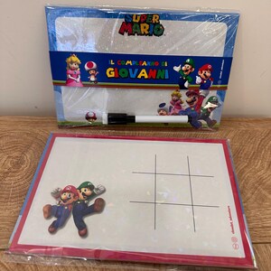 Puede incluir: Dos pizarras blancas de borrado en seco con tem&aacute;tica de Super Mario en envoltorio de pl&aacute;stico transparente. La pizarra superior incluye el logo de Super Mario, personajes y el texto "IL COMPLEANNO DI GIOVANNI" con un marcador negro. La inferior muestra a Mario y Luigi.