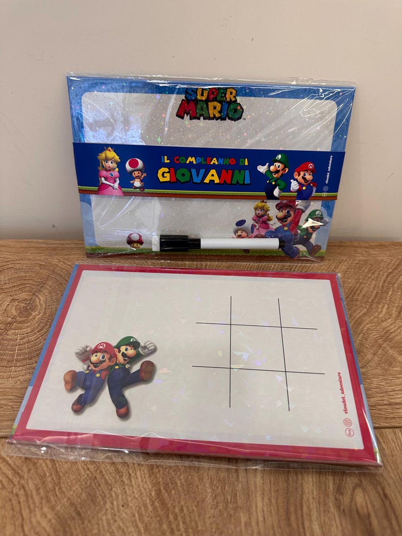 Puede incluir: Dos pizarras blancas de borrado en seco con tem&aacute;tica de Super Mario en envoltorio de pl&aacute;stico transparente. La pizarra superior incluye el logo de Super Mario, personajes y el texto "IL COMPLEANNO DI GIOVANNI" con un marcador negro. La inferior muestra a Mario y Luigi.