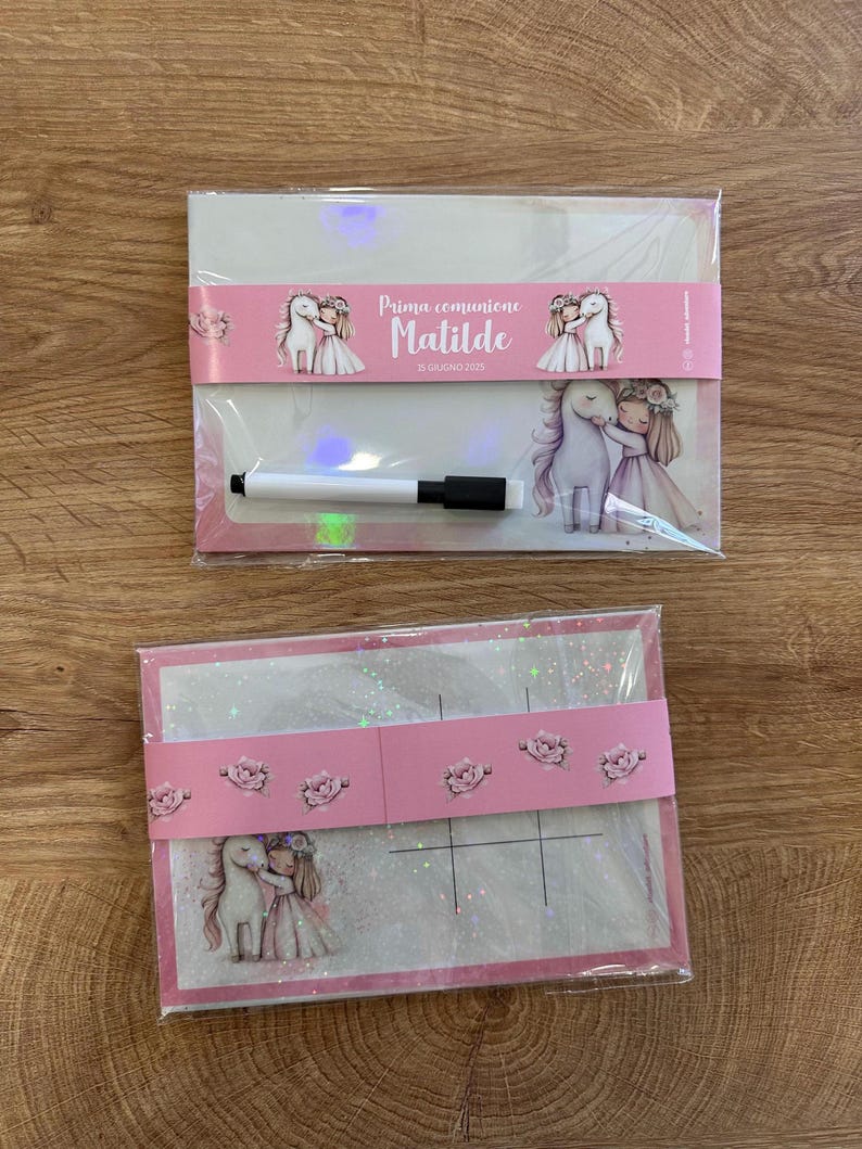 Puede incluir: Dos pizarras blancas rectangulares envueltas en pl&aacute;stico transparente. La superior tiene una banda rosa con el texto "Prima comunione Matilde 15 GIUGNO 2025" y un marcador blanco. Ambas muestran un gr&aacute;fico de una ni&ntilde;a con un unicornio.