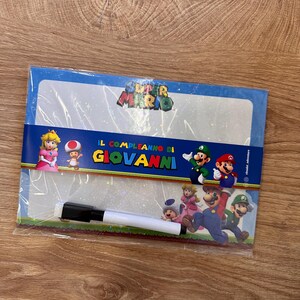 Puede incluir: Una pizarra blanca de borrado en seco con tem&aacute;tica de Super Mario en un envoltorio de pl&aacute;stico transparente. La pizarra muestra a la Princesa Peach, Toad, Mario y Luigi, con el texto "IL COMPLEANNO DI GIOVANNI" impreso en el centro. Incluye un rotulador negro.