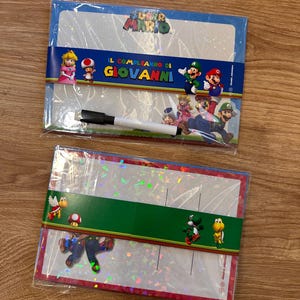 Puede incluir: Dos pizarras blancas rectangulares con tem&aacute;tica de Super Mario en envoltorio de pl&aacute;stico transparente. La superior muestra a la Princesa Peach, Toad, Luigi y Mario con el texto IL COMPLEANNO DI GIOVANNI y un rotulador negro. La inferior muestra a Yoshi y Koopa.
